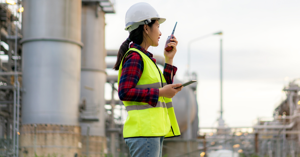 A woman in a green safety vest and white hard hat standing outside while using a two-way radio at an oil refinery.