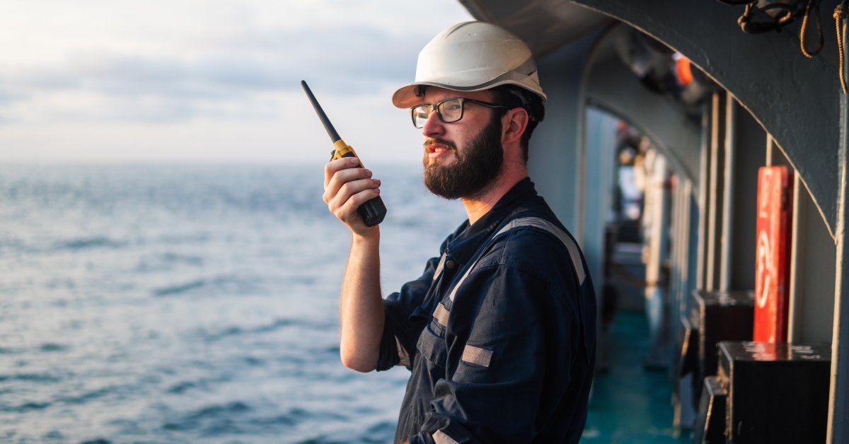 A man in a blue reflective uniform and a white hard hat using a two-way radio while standing on the deck of a ship.